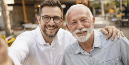 Father and Son: A warm and touching moment between a father and his adult son is captured. Both are smiling at the camera, showcasing their close bond. They are outdoors, likely in a casual café or restaurant setting, as suggested by the background. The father, with a white beard and hair, is wearing a light denim shirt, while the son, wearing glasses and a white shirt, has his arm around his father's shoulder.