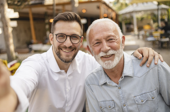 Father and Son: A warm and touching moment between a father and his adult son is captured. Both are smiling at the camera, showcasing their close bond. They are outdoors, likely in a casual café or restaurant setting, as suggested by the background. The father, with a white beard and hair, is wearing a light denim shirt, while the son, wearing glasses and a white shirt, has his arm around his father's shoulder.