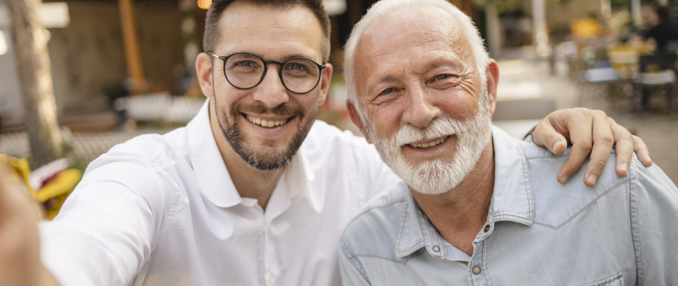 Father and Son: A warm and touching moment between a father and his adult son is captured. Both are smiling at the camera, showcasing their close bond. They are outdoors, likely in a casual café or restaurant setting, as suggested by the background. The father, with a white beard and hair, is wearing a light denim shirt, while the son, wearing glasses and a white shirt, has his arm around his father's shoulder.